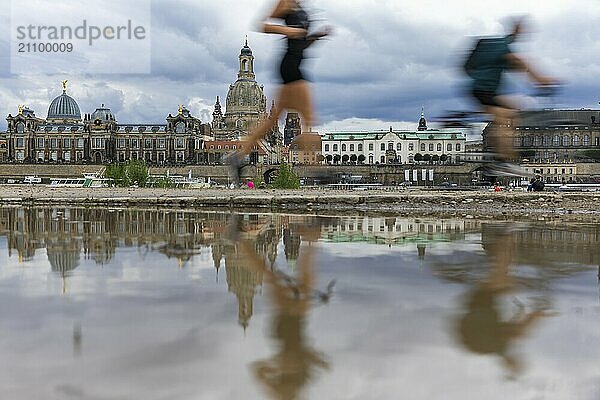 Die Dresdner Silhouette mit einem Radfahrer und einer Joggerin und dunklen Wolken hinter der Frauenkirche  spiegelt sich am Elberadweg in einer Pfütze.  Elberadweg  Dresden  Sachsen  Deutschland  Europa