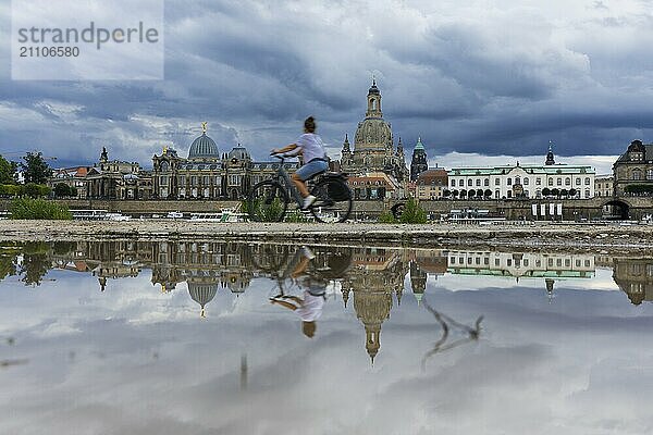 Die Dresdner Silhouette mit einem Radfahrer und dunklen Wolken hinter der Frauenkirche  spiegelt sich am Elberadweg in einer Pfütze.  Elberadweg  Dresden  Sachsen  Deutschland  Europa