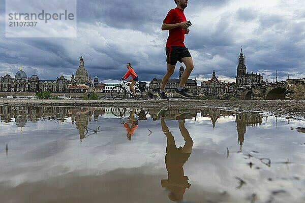 Die Dresdner Silhouette mit einem Radfahrer und einem Jogger und dunklen Wolken hinter der Frauenkirche  spiegelt sich am Elberadweg in einer Pfütze.  Elberadweg  Dresden  Sachsen  Deutschland  Europa