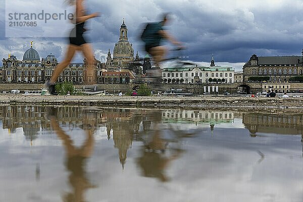 Die Dresdner Silhouette mit einem Radfahrer und einer Joggerin und dunklen Wolken hinter der Frauenkirche  spiegelt sich am Elberadweg in einer Pfütze.  Elberadweg  Dresden  Sachsen  Deutschland  Europa