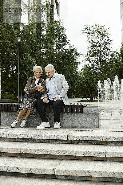 Senior woman using smart phone and sitting with man near staircase at street