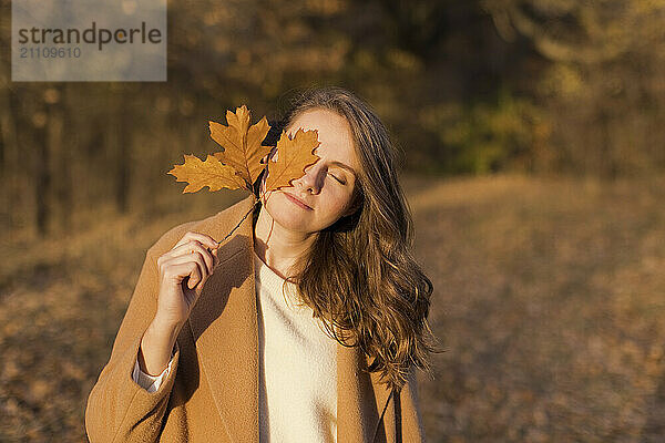 Woman covering eye with autumn leaf at forest