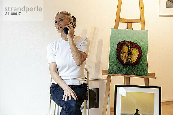 Artist talking on smart phone and sitting near paintings in art studio