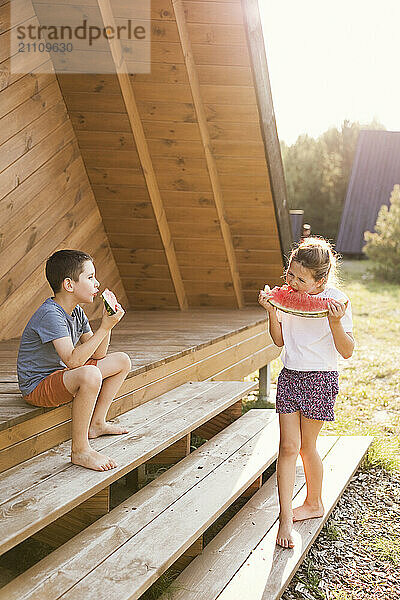 Siblings eating watermelon on porch of wooden house