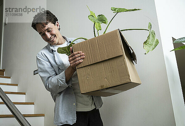 Smiling man carrying cardboard box with houseplant and moving down stairs at new home