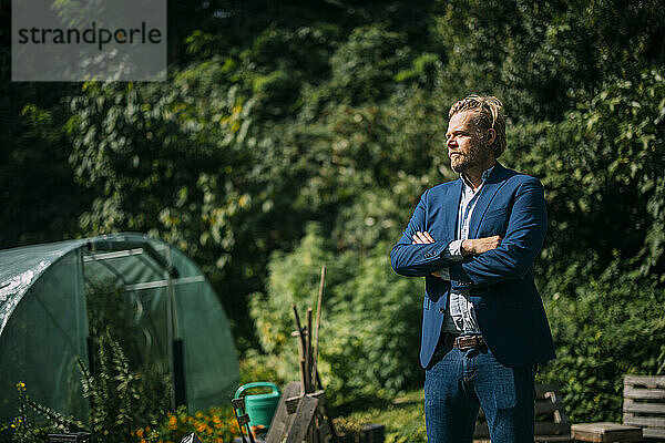 Mature businessman standing with arms crossed outside greenhouse at sunny day