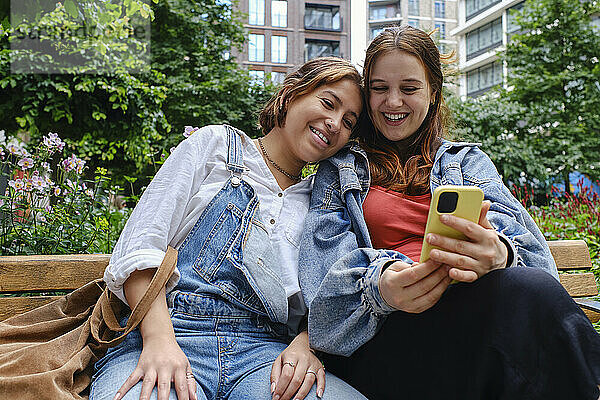 Young woman with girlfriend using smart phone and sitting on bench at park