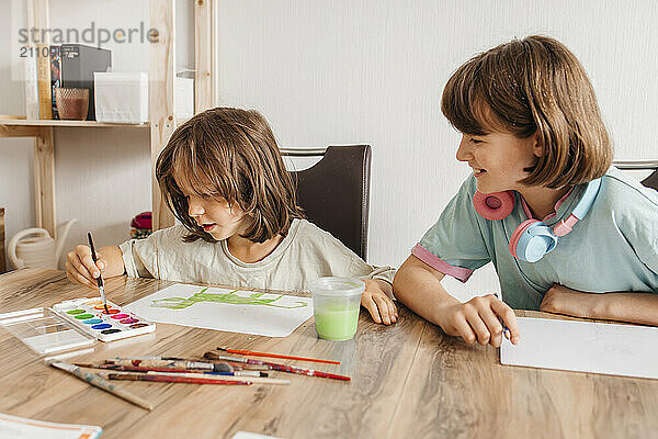 Smiling girl sitting near table with brother painting on paper at home