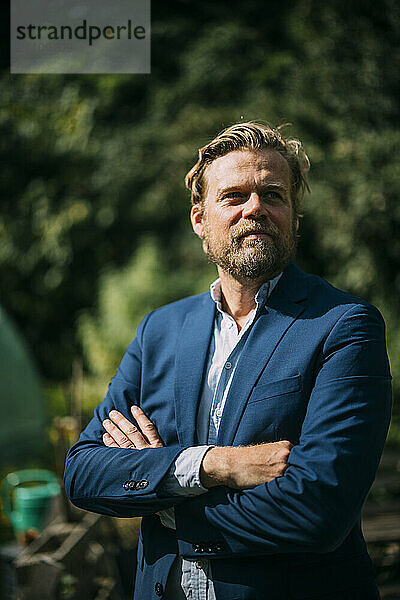 Smiling businessman wearing blazer and standing with arms crossed at sunny day