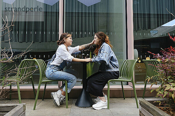 Gay woman sitting near table and pulling cheek of girlfriend at outdoor restaurant