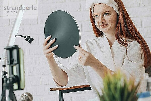 Teenage girl in bathrobe applying makeup in front of a mirror  recording a beauty vlog at home.