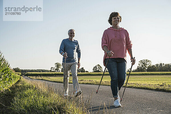 Senior couple holding hiking poles and walking on road near field