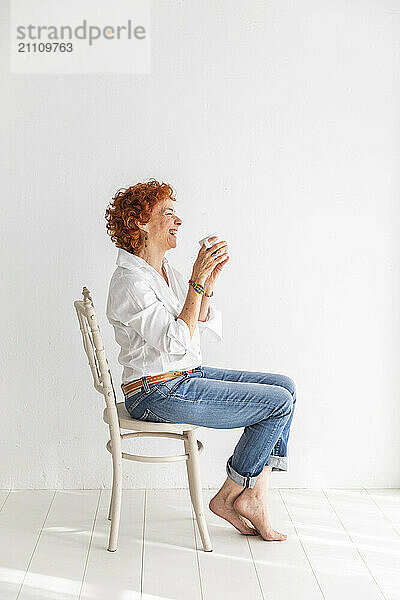 Happy woman holding coffee mug and sitting on chair near white wall