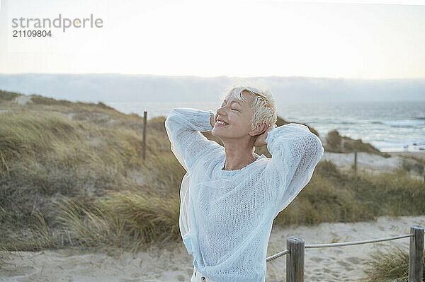 Happy mature woman with hands behind head standing at beach