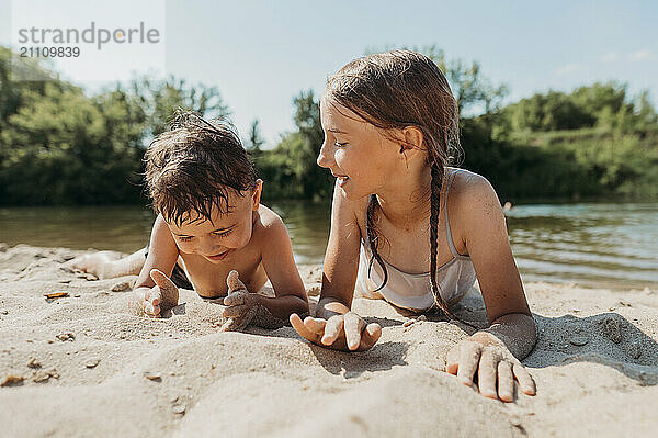 Girl playing with brother and lying on sand near river