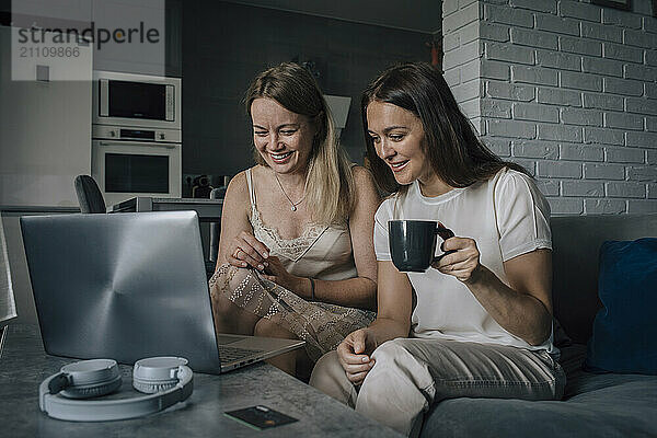 Happy women watching laptop and holding cup at home