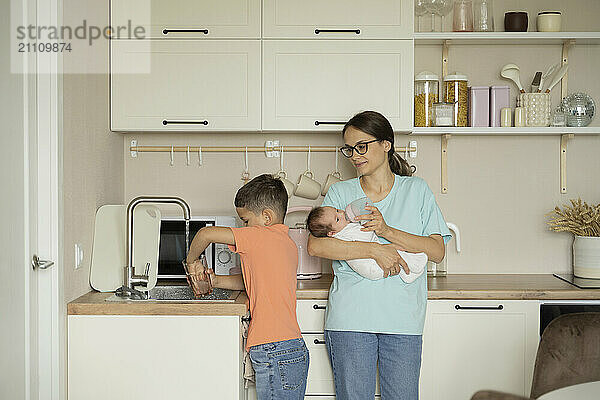 Mother feeding baby and son washing glass in kitchen