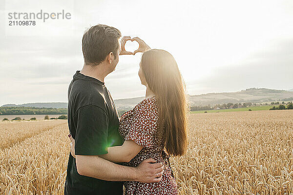 Young couple making heart shape and standing in agricultural field at sunset