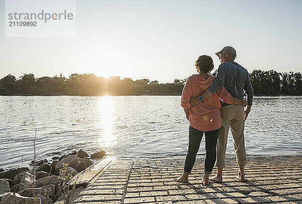 Affectionate senior couple standing with arm around and looking at sunset near river