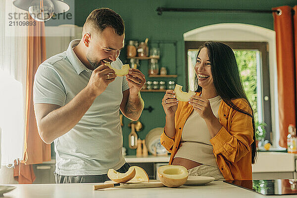 Smiling man with pregnant wife eating sliced melon in kitchen