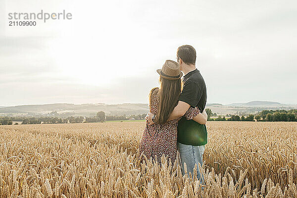 Young couple standing with arms around and looking at view in agricultural field