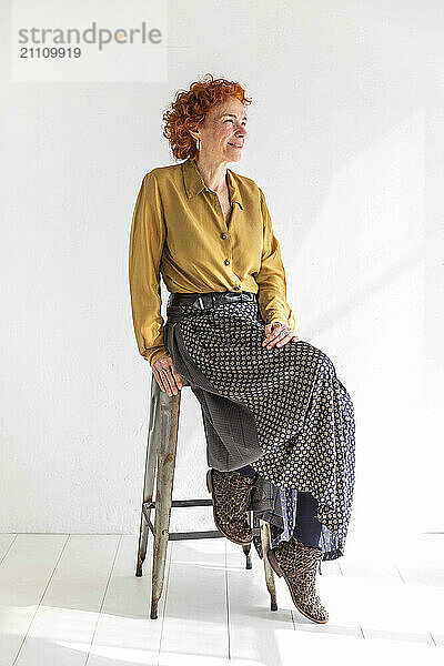 Thoughtful redhead woman sitting on stool in front of white wall