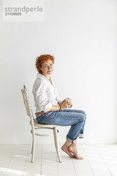 Redhead woman holding coffee mug and sitting on chair in front of white wall