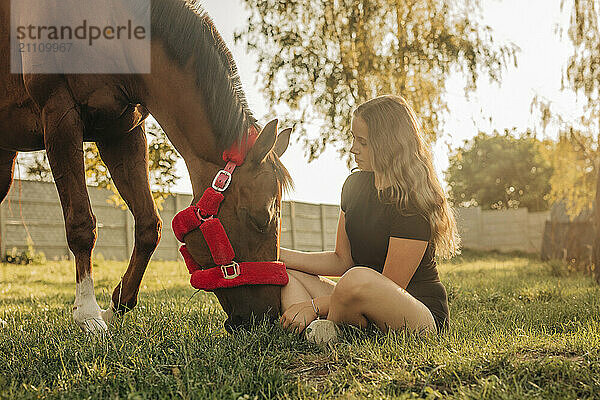 Girl sitting on grass with horse at farm