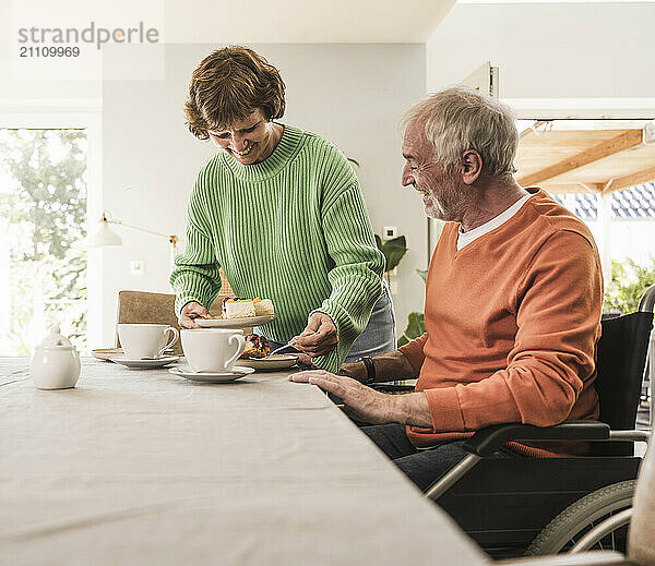 Smiling woman serving slice of cake to husband on wheelchair at home