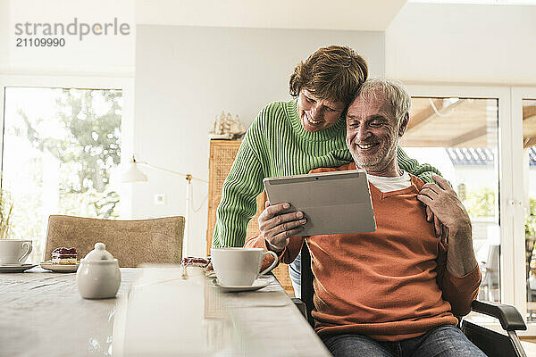 Affectionate woman with husband on wheelchair using tablet PC near table at home