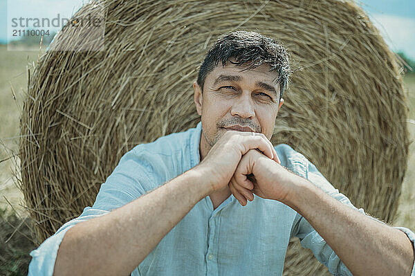 Mature man with hands on chin near hay bale in field