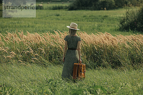 Woman wearing hat and holding suitcase standing near reeds