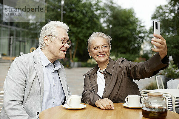 Happy woman holding smart phone and taking selfie with man at cafe