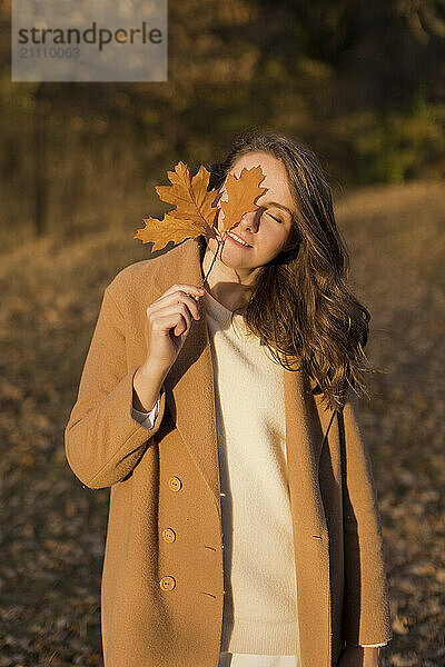 Woman wearing beige coat covering eye with autumn leaf at forest