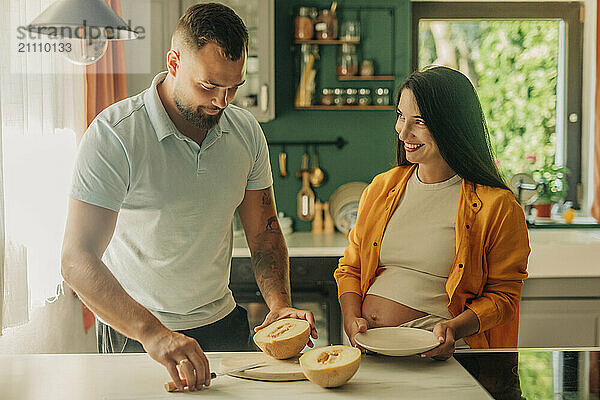 Pregnant woman with husband cutting muskmelon in kitchen