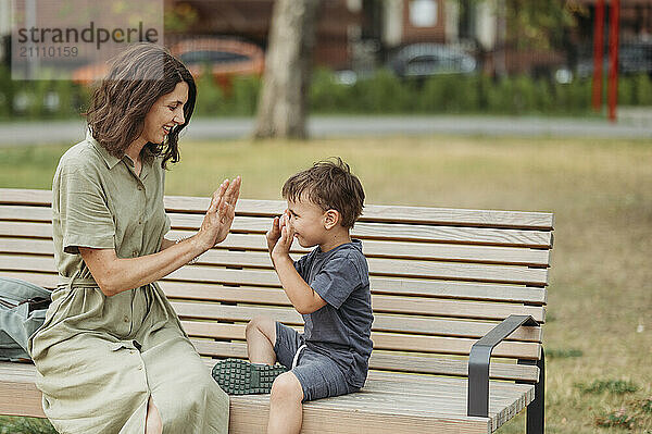 Smiling mother playing clapping game with son sitting on bench at playground