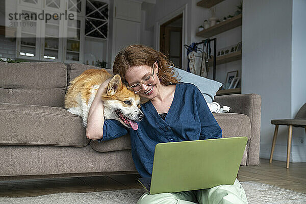 Smiling woman with laptop embracing pet dog at home