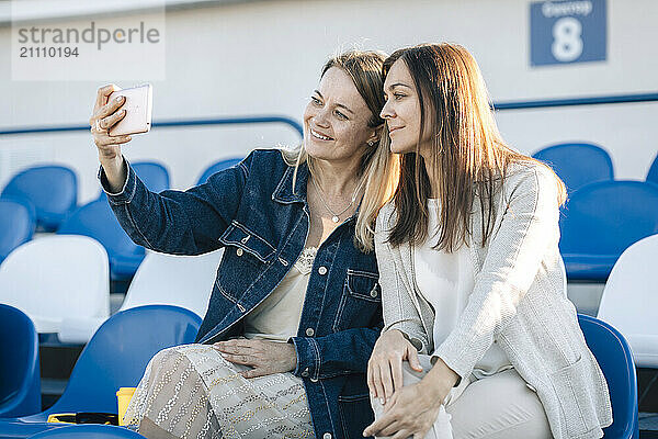 Friends taking selfie through smart phone sitting on chairs at stadium