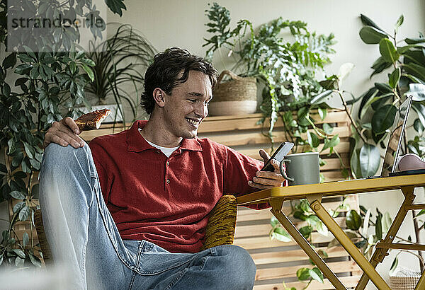 Smiling man using smart phone and holding pizza slice sitting on chair at home