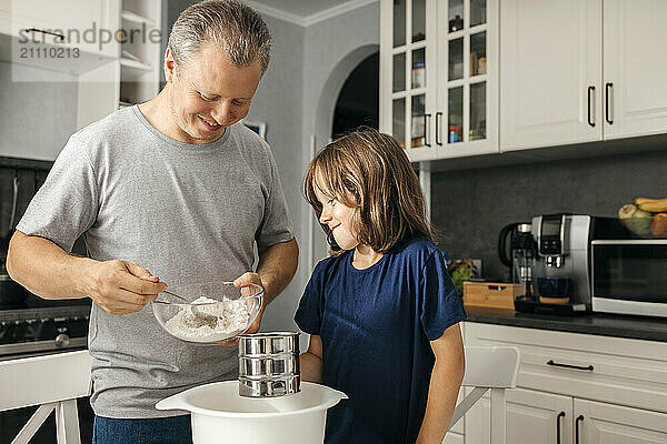 Smiling man pouring flour with son holding sifter in kitchen