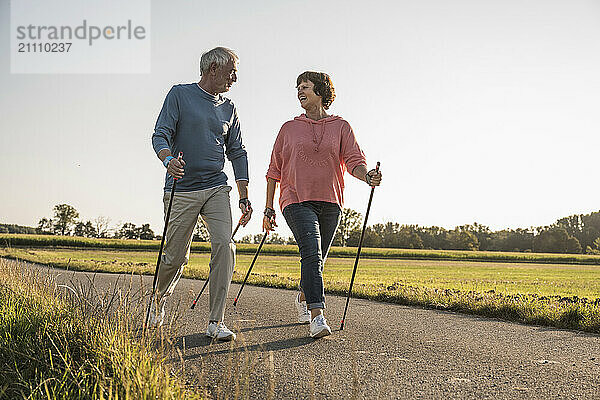 Senior couple walking with hiking poles on road near field