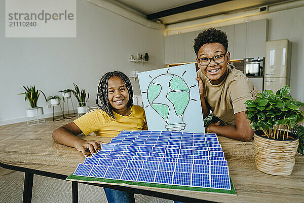 Happy siblings sitting with solar panels project and light bulb drawing at table