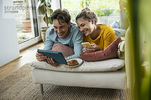 Smiling woman lying on sofa with boyfriend using tablet PC at home