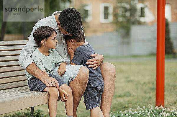 Father consoling upset son sitting on bench at park