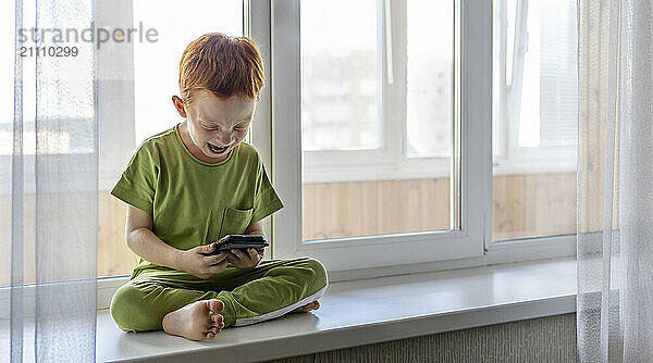 Redhead boy with smart phone laughing and sitting on window sill at home