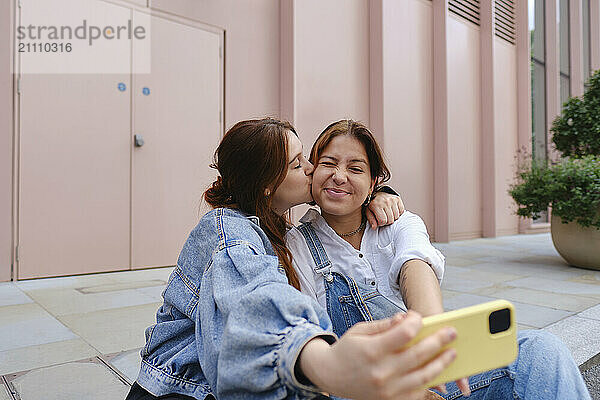 Young woman kissing girlfriend and taking selfie on sidewalk