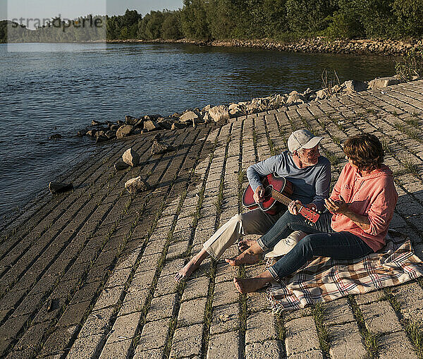 Senior man playing guitar and sitting with woman on picnic blanket near river