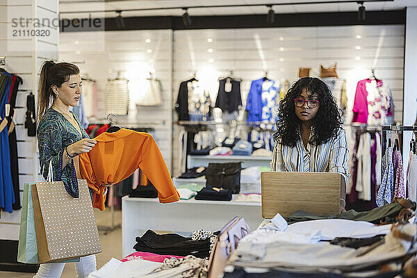 Woman holding dress and walking towards friend using laptop at fashion store