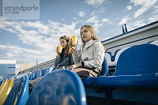 Girls holding disposable cups and sitting on chairs at stadium