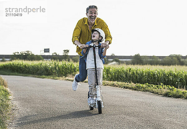 Cheerful grandfather enjoying riding push scooter with grandson on street